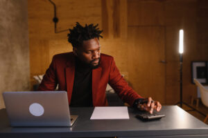 A businessman in a red suit jacket sits in his at-home office at his computer, analyzing the stock market using a calculator.