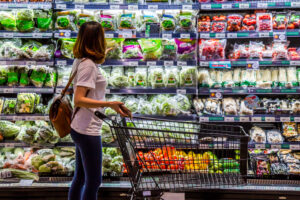 A woman with an empty cart shops along the vegetable aisle of a grocery store.