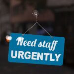 A blue sign on a shop windowsill that reads “Need Staff Urgently.”