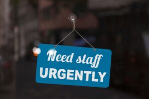 A blue sign on a shop windowsill that reads “Need Staff Urgently.”