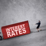 Two people with a rope dragging a brick that reads “interest rates.”