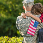 A father holding his son while in uniform with American flags present.