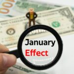 A magnifying glass reading “January effect” with a figurine standing over a dollar-bill.