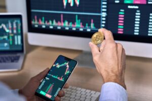 A man holding a Bitcoin in one hand and a phone with a stock chart in the other in front of a computer screen.