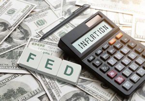 White tiles that read “FED” lie on piles of hundred-dollar bills. To the right, a calculator reads “INFLATION” with a pen above it.