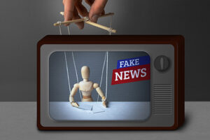 A wooden figurine on TV being controlled by a puppeteer while the screen reads “FAKE NEWS.”