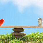 A scale with a heart on the left and a rolled-up stack of money on the right in a serene grass field with blue skies.