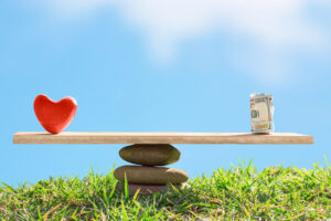 A scale with a heart on the left and a rolled-up stack of money on the right in a serene grass field with blue skies.