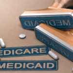 Two rubber stamps with the words “MEDICARE” and “MEDICAID” printed over a paper background with loose pills lying around.
