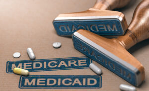 Two rubber stamps with the words “MEDICARE” and “MEDICAID” printed over a paper background with loose pills lying around.