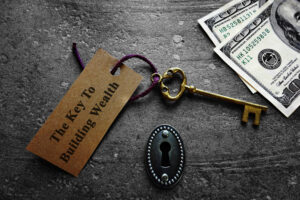 A skeleton key with a paper attached that reads “The key to building wealth” next to hundred-dollar bills.