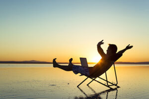 A man sitting on a chair on a beach with his hands and legs in the air, while holding a laptop.