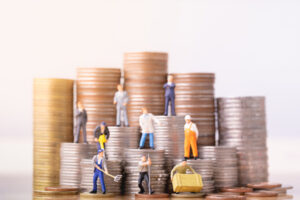 Miniature people standing on piles of money.