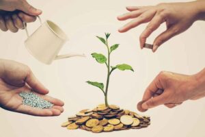 Several hands helping a small tree on top of a pile of money to grow.